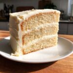 Close-up of a tall, three-layer slice of vanilla cake recipe with thick white vanilla buttercream frosting.