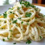 A close-up of creamy spaghetti coated in a rich sauce, garnished with fresh parsley and black pepper, featuring garlic parmesan flavor.