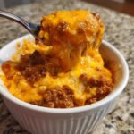 A spoonful of hot chili cheese dip being lifted from a white bowl, showing melted, stringy cheddar cheese.