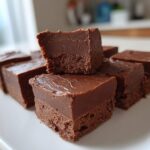 Several squares of rich, dark chocolate fudge stacked on a white plate, showing the smooth texture of the fudge.
