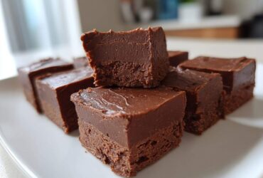 Several squares of rich, dark chocolate fudge stacked on a white plate, showing the smooth texture of the fudge.