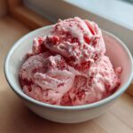 Three generous scoops of pink strawberry ice cream with visible fruit pieces served in a light ceramic bowl.