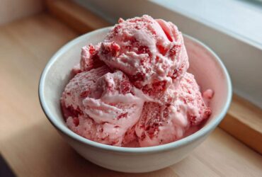 Three generous scoops of pink strawberry ice cream with visible fruit pieces served in a light ceramic bowl.