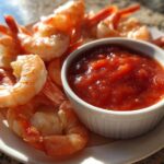 Close-up of cooked, chilled shrimp arranged around a small white bowl of bright red shrimp cocktail sauce.