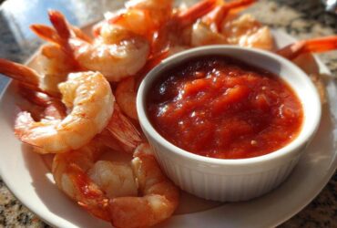 Close-up of cooked, chilled shrimp arranged around a small white bowl of bright red shrimp cocktail sauce.