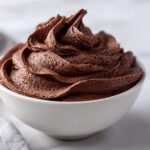 A close-up of rich, dark chocolate frosting swirled high in a small white bowl.