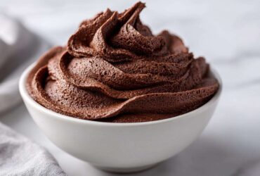 A close-up of rich, dark chocolate frosting swirled high in a small white bowl.