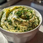 Close-up of a creamy, pale yellow olive dip swirled in a white bowl and topped with fresh chopped parsley.