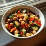 A close-up view of a vibrant bean salad featuring chickpeas, black beans, cucumber, and red pepper in a white bowl.