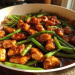 Close-up of bite-sized chicken pieces coated in a dark glaze mixed with bright green beans in a skillet.