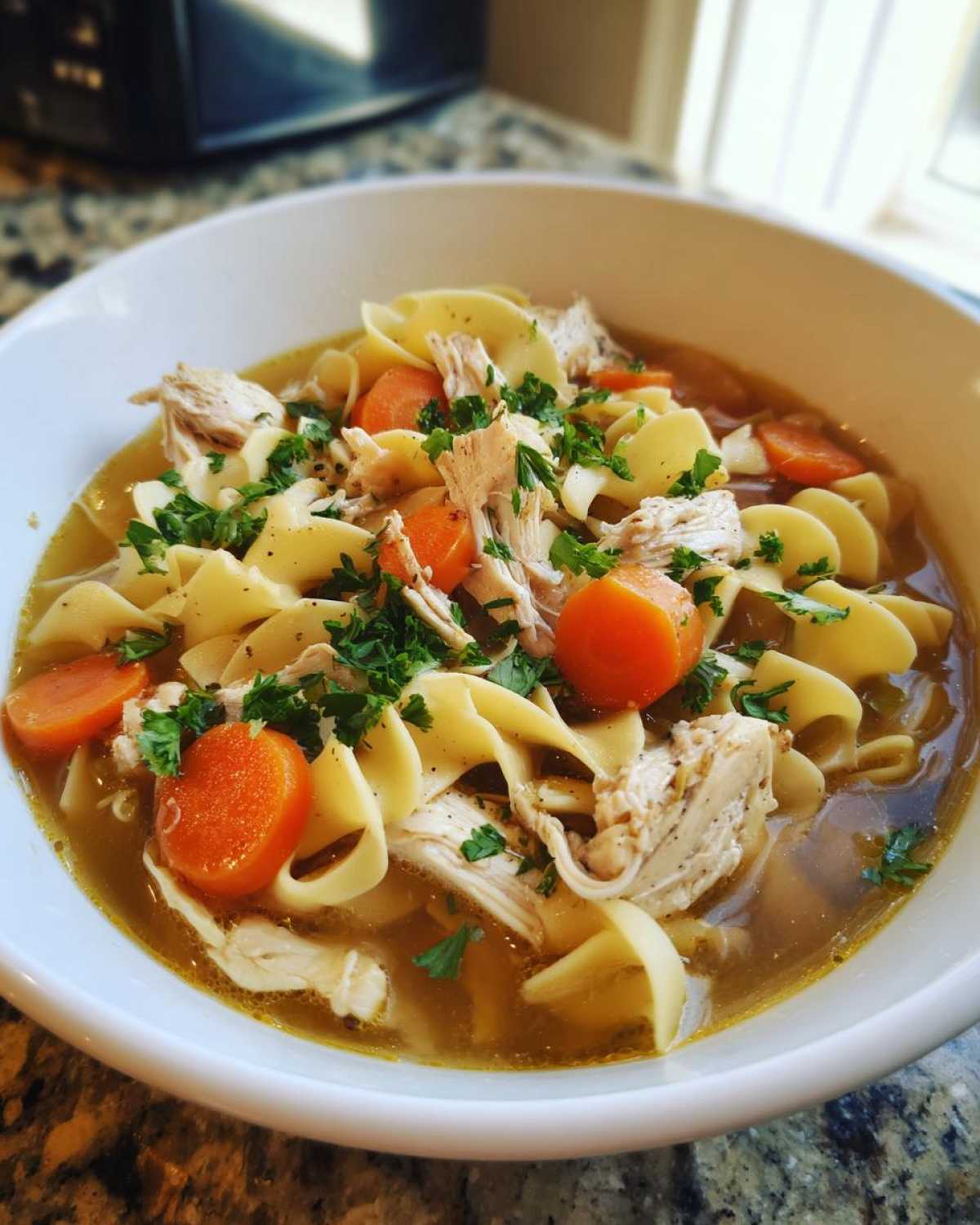 A close-up view of a white bowl filled with homemade chicken noodle soup, featuring egg noodles, shredded chicken, and sliced carrots.