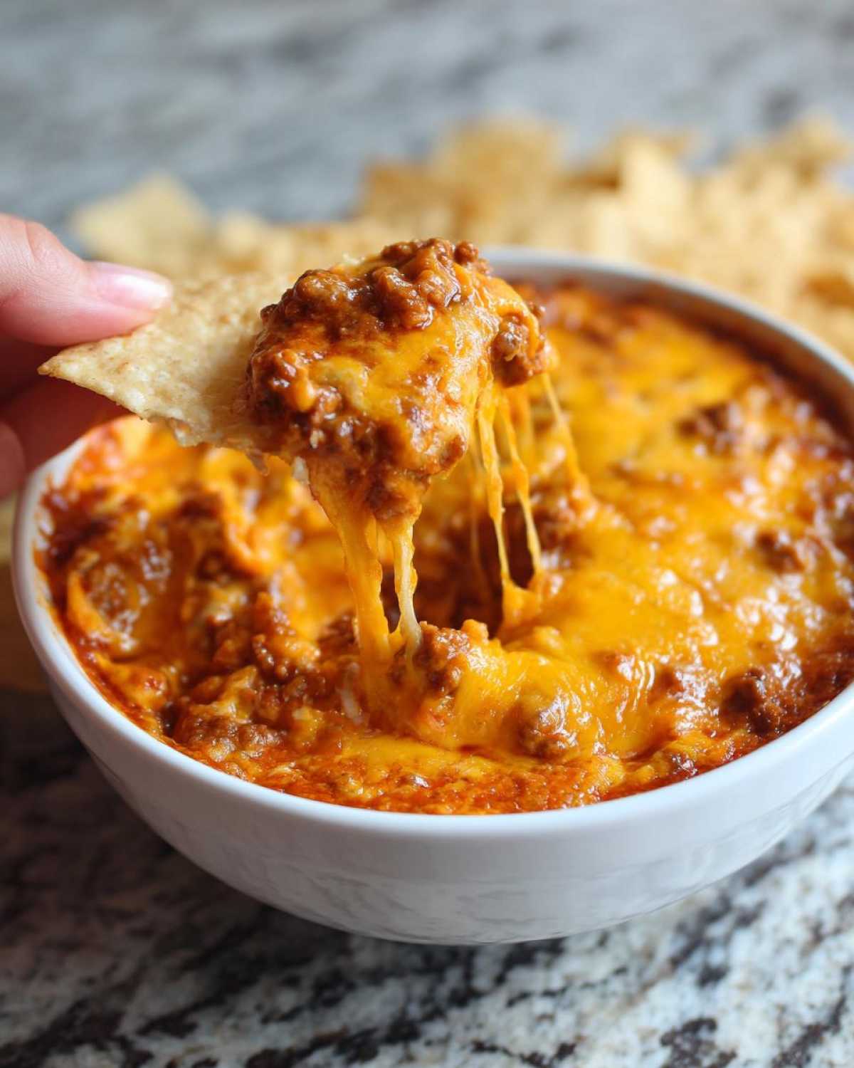 A tortilla chip scooping up a generous portion of hot chili cheese dip, showing a dramatic cheese pull.