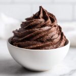 A close-up of rich, dark chocolate frosting swirled high in a small white bowl.
