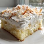A moist slice of coconut poke cake topped with whipped cream and toasted coconut flakes.