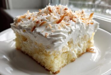 A moist slice of coconut poke cake topped with whipped cream and toasted coconut flakes.