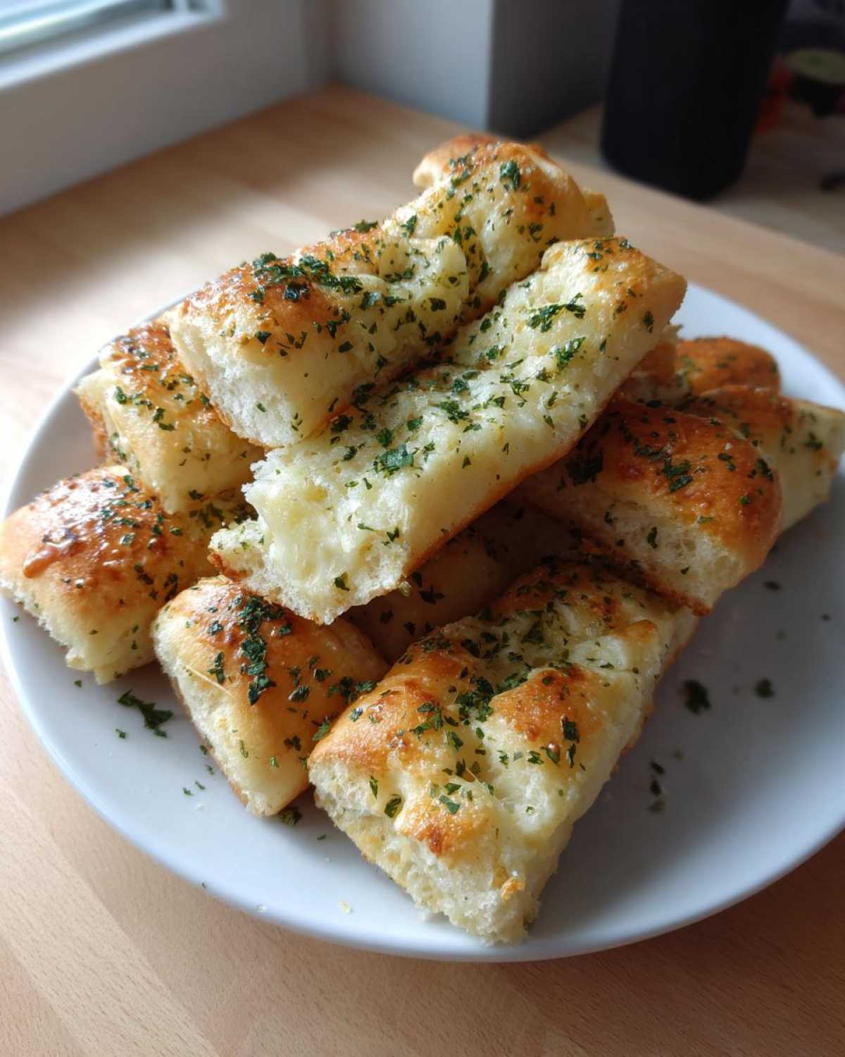 A stack of freshly baked, soft garlic breadsticks sprinkled generously with chopped green parsley.
