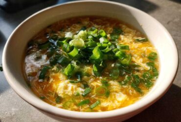 A close-up bowl of hot egg drop soup, featuring wispy cooked egg ribbons garnished heavily with fresh green scallions.