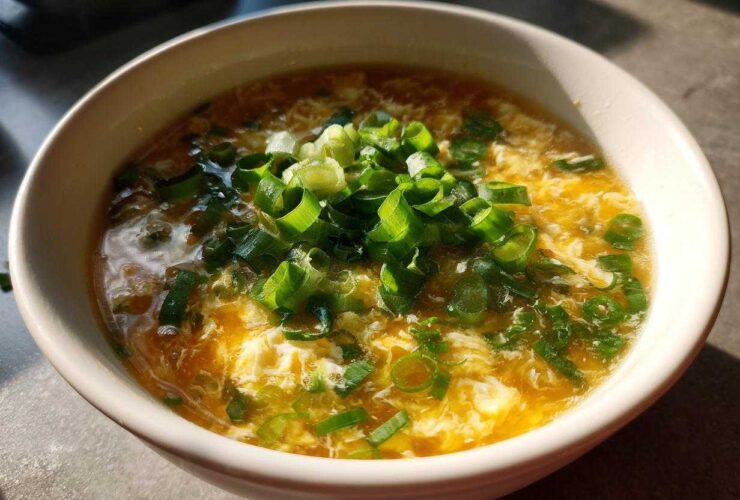 A close-up bowl of hot egg drop soup, featuring wispy cooked egg ribbons garnished heavily with fresh green scallions.