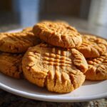 A close-up shot of several delicious peanut butter cookies stacked on a white plate, showing the classic crisscross fork pattern.
