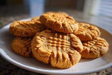 A close-up shot of several delicious peanut butter cookies stacked on a white plate, showing the classic crisscross fork pattern.