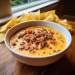 A white bowl filled with creamy, cheesy rotel dip topped with ground beef and diced tomatoes, served with tortilla chips.