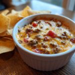 Close-up of a creamy, cheesy rotel dip topped with ground beef and tomatoes, served next to tortilla chips.