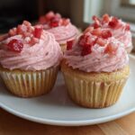 Four amazing strawberry cupcakes with swirled pink frosting and diced strawberries on top, served on a white plate.