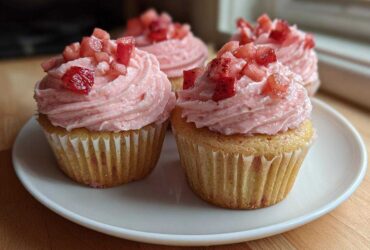 Four amazing strawberry cupcakes with swirled pink frosting and diced strawberries on top, served on a white plate.