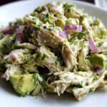 Close-up of shredded chicken mixed with creamy avocado dressing, chunks of avocado, and slices of red onion in an avocado chicken salad.