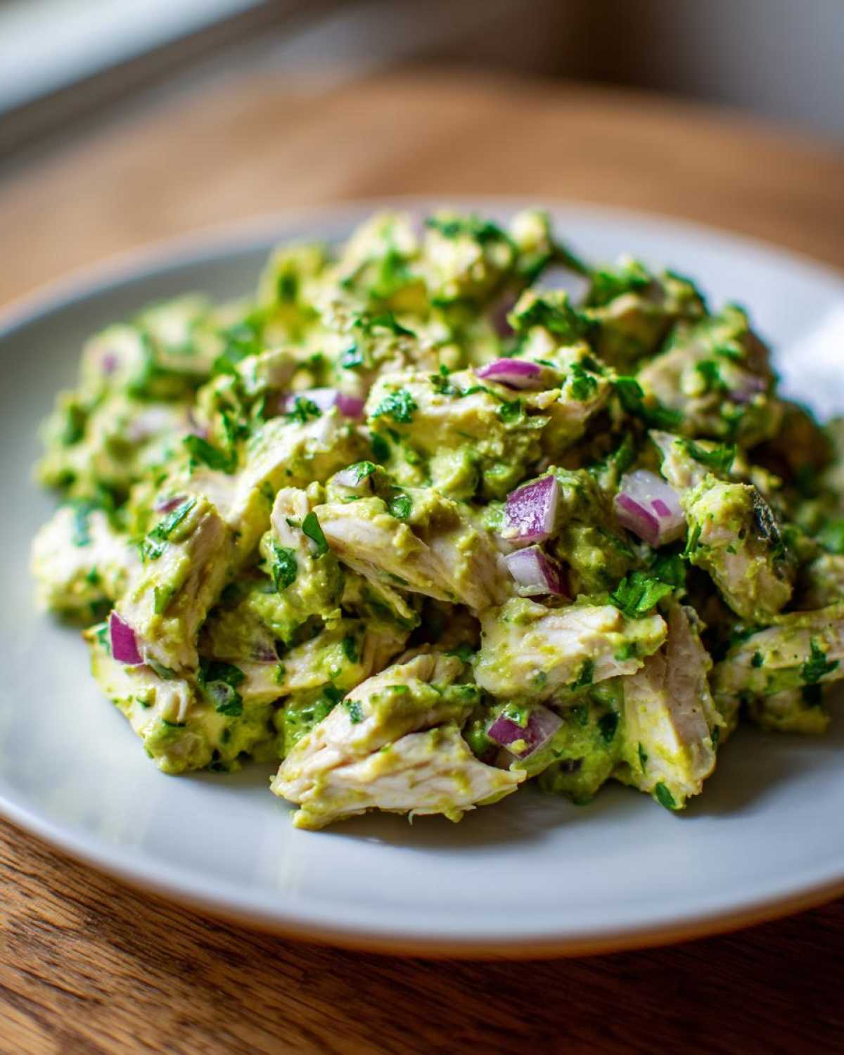 A close-up of creamy avocado chicken salad mixed with chunks of red onion and cilantro on a light gray plate.