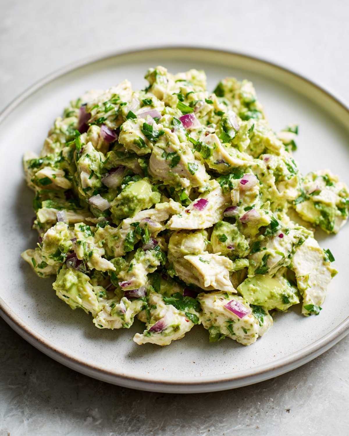 A mound of fresh avocado chicken salad mixed with red onion and herbs served on a light gray plate.