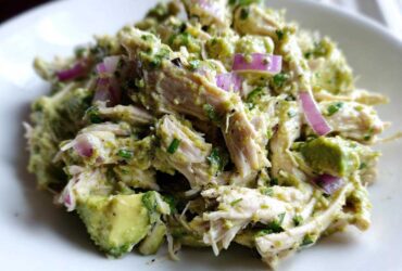 Close-up of shredded chicken mixed with creamy avocado dressing, chunks of avocado, and slices of red onion in an avocado chicken salad.