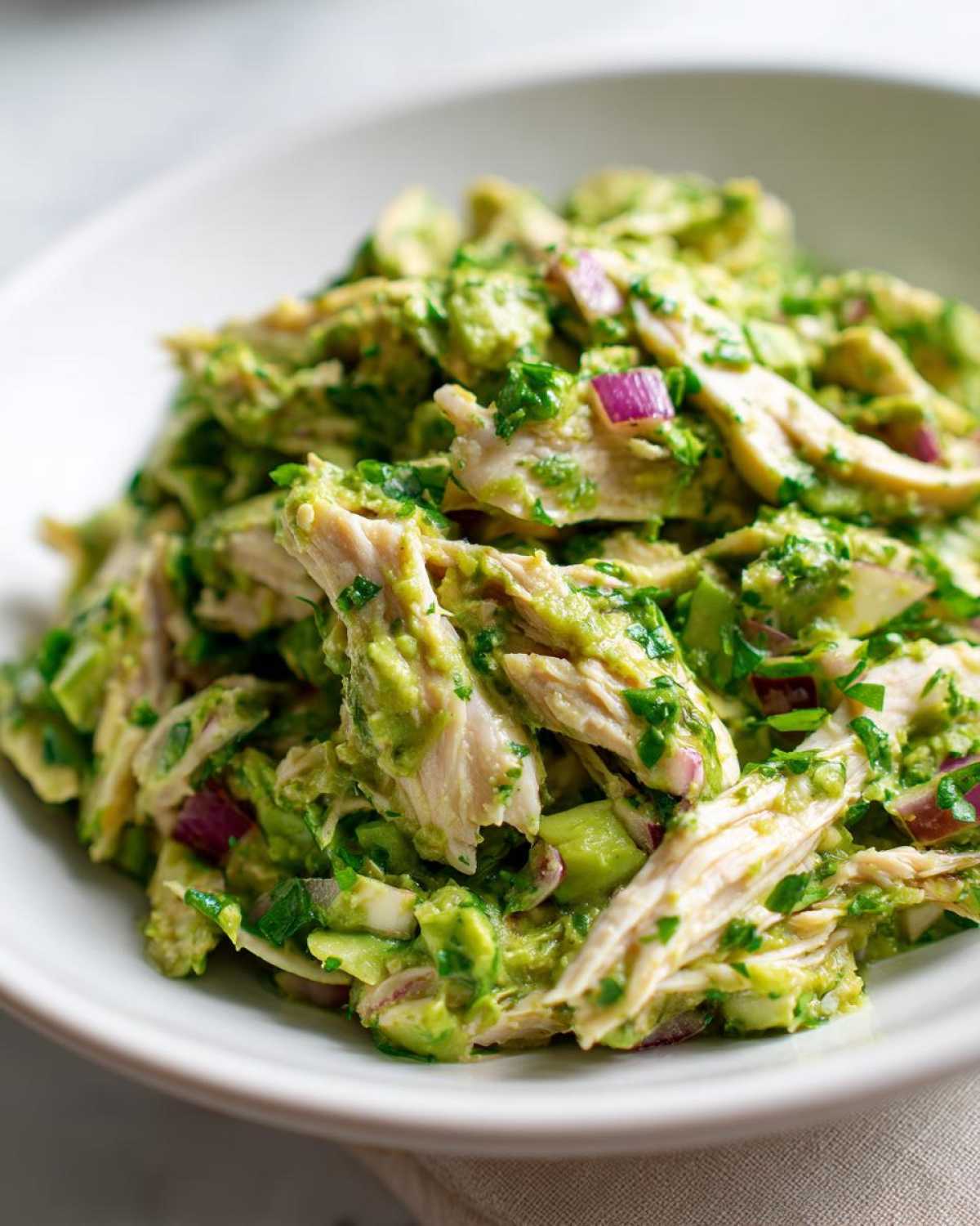 Close-up of shredded chicken mixed with creamy avocado dressing, red onion, and herbs in an avocado chicken salad.