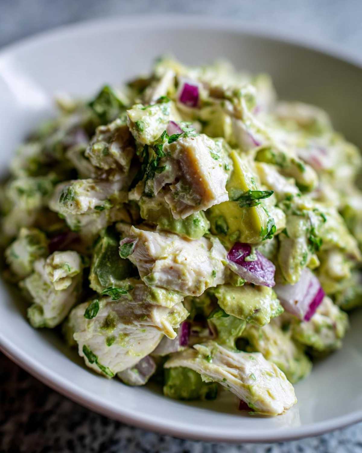 Close-up shot of chunks of chicken mixed with creamy avocado dressing, red onion, and herbs in a white bowl, featuring avocado chicken salad.