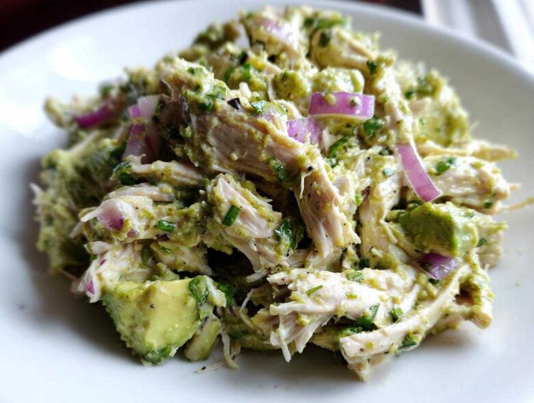 Close-up of shredded chicken mixed with creamy avocado dressing, chunks of avocado, and slices of red onion in an avocado chicken salad.