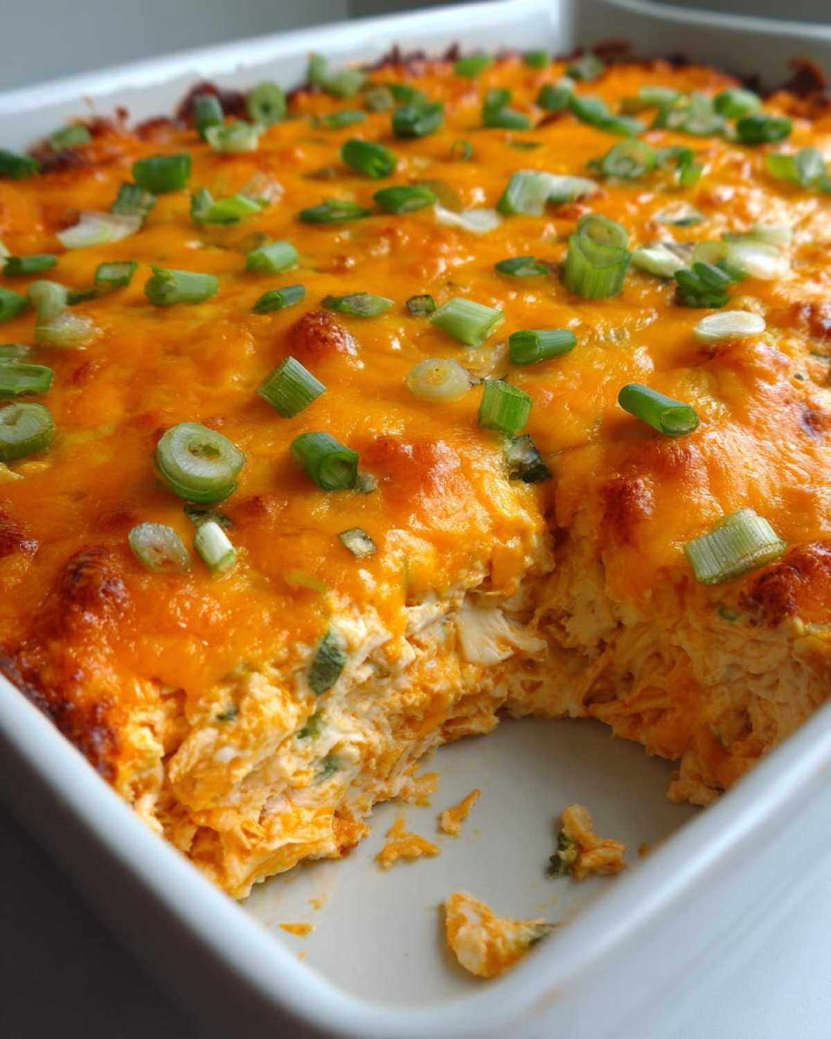 Close-up of baked Buffalo chicken dip in a white dish, showing melted cheddar cheese and chopped green onions.