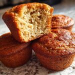 Close-up of four golden brown banana protein muffins, one cut in half showing the moist interior texture.