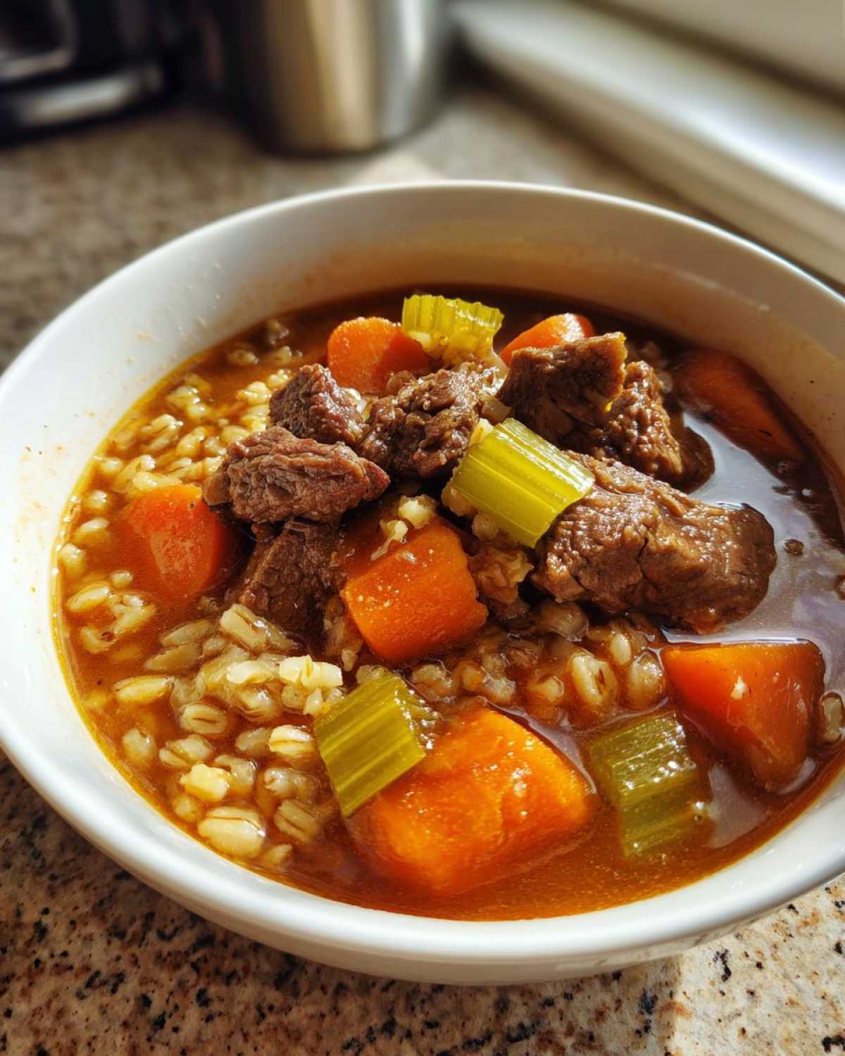 Close-up of a white bowl filled with rich beef barley soup, featuring chunks of beef, carrots, celery, and barley.
