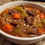 Close-up of a white bowl filled with rich beef barley soup, featuring chunks of beef, carrots, celery, and barley.