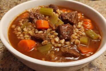Close-up of a white bowl filled with rich beef barley soup, featuring chunks of beef, carrots, celery, and barley.