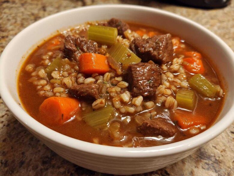 Close-up of a white bowl filled with rich beef barley soup, featuring chunks of beef, carrots, celery, and barley.