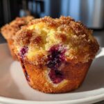 A close-up of a golden brown blueberry muffin featuring a thick, sugary crumb topping and visible burst blueberries.