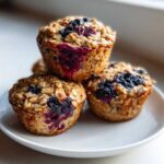Three freshly baked blueberry oatmeal muffins stacked on a small white plate, showing visible oats and burst blueberries.