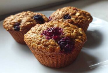 Close-up of three golden brown blueberry oatmeal muffins topped with visible oats and bursting blueberries on a white plate.