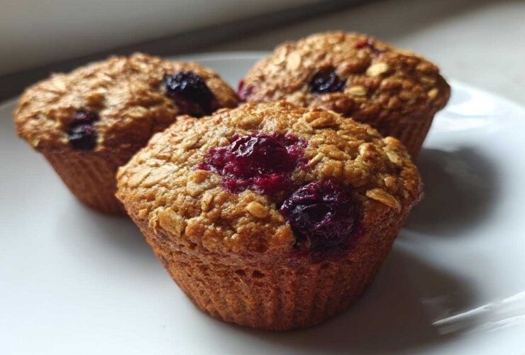 Close-up of three golden brown blueberry oatmeal muffins topped with visible oats and bursting blueberries on a white plate.