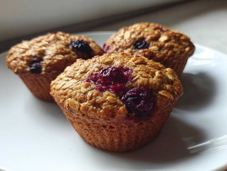 Close-up of three golden brown blueberry oatmeal muffins topped with visible oats and bursting blueberries on a white plate.