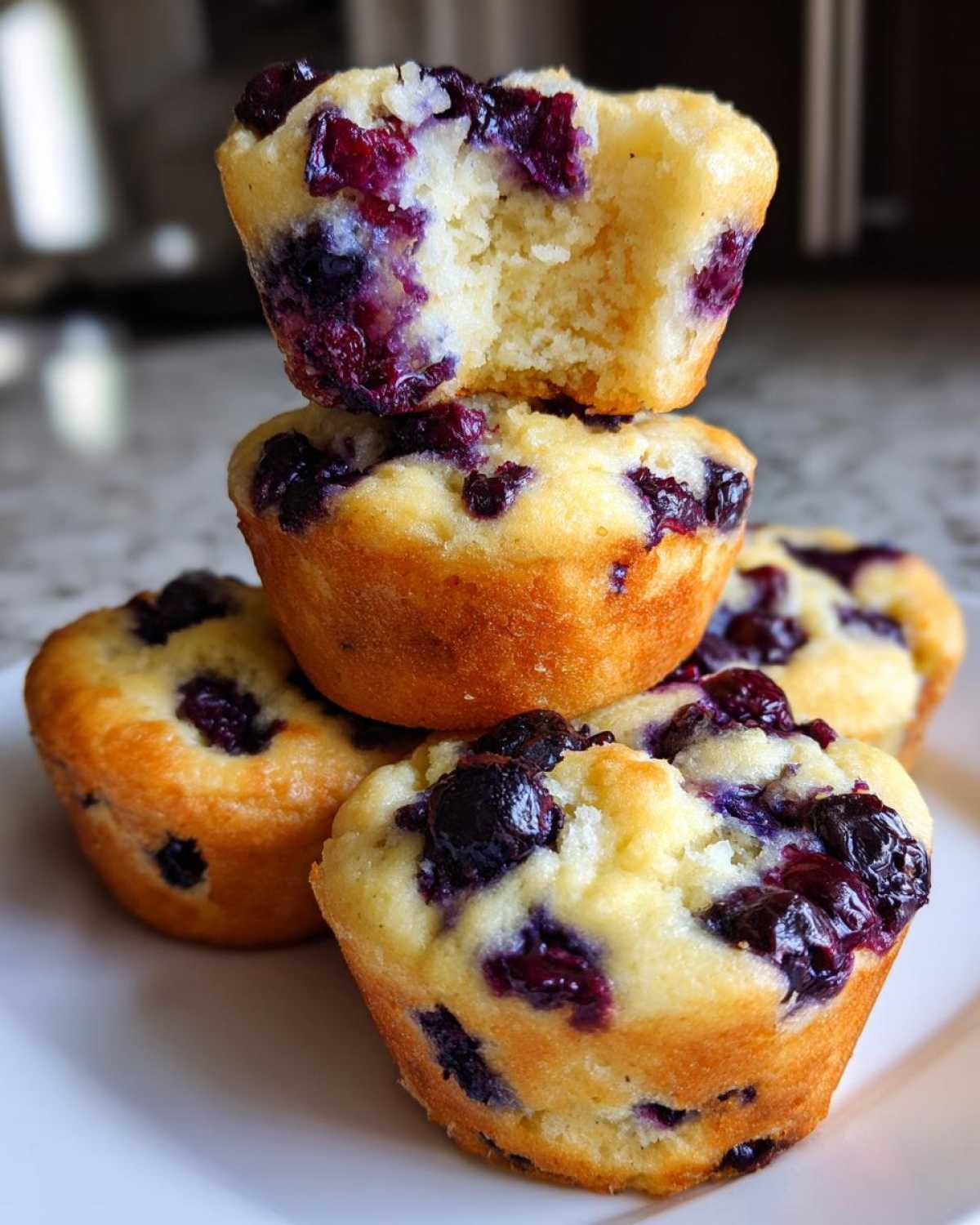 A stack of golden-brown blueberry protein muffins, with the top one broken open to show the moist interior.