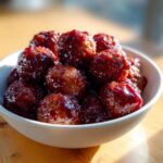 A white bowl overflowing with glossy, saucy grape jelly meatballs, sitting on a wooden surface.