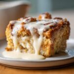 Close-up of a square serving of rich bread pudding topped with thick vanilla glaze dripping down the sides.