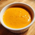 Close-up of smooth, bright orange nacho cheese sauce served in a small white ramekin on a wooden surface.